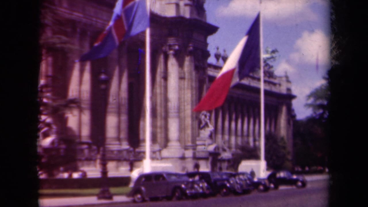 1939 Paris 8mm frame showing national flags before a neoclassical building, with classic cars parked at the curb.