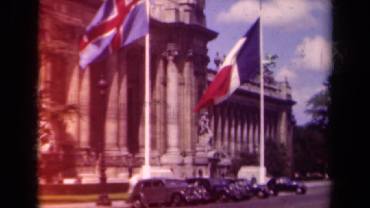 1939 Paris frame with Icelandic and Dutch flags visible before an ornate stone building and parked period automobiles.