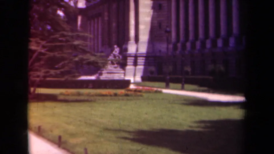 1939 Paris home-movie frame with a statue, green lawn, walkway, trees, and the columned stone facade in view.