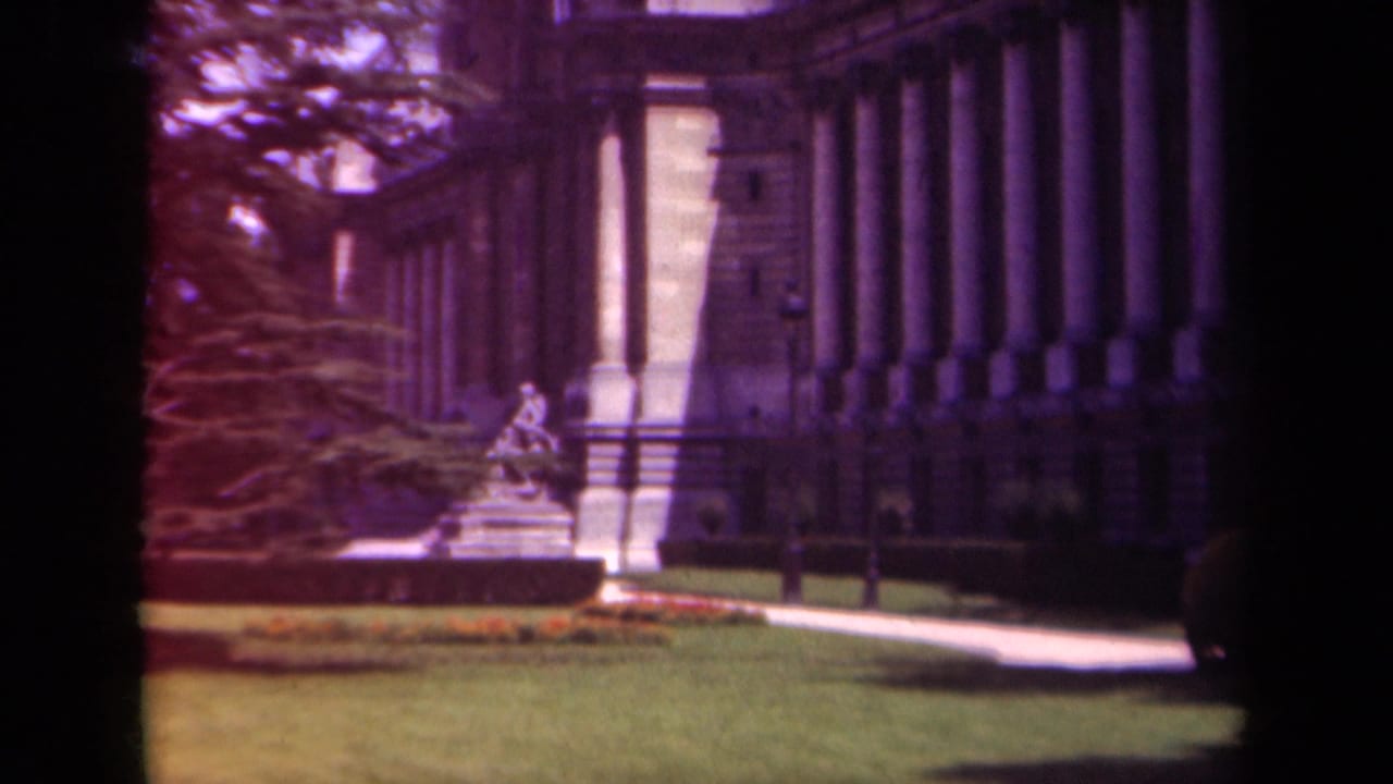 Soft 8mm view of a Paris garden with manicured grass, flowerbeds, trees, and a classical building with tall columns behind it.