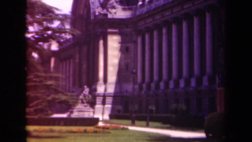 1939 Paris frame with a classical colonnade, shaded facade, garden sculpture, lawn, flowerbed, and tree branches in soft color film.