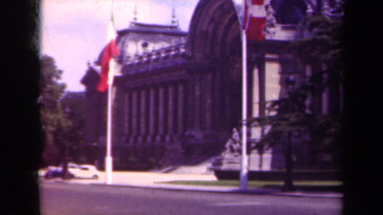 1939 Paris view of an ornate facade with flags near the curb and a light-colored period car on the street.