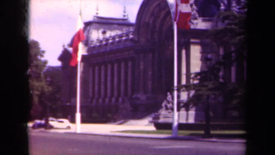 1939 Paris view of an ornate facade with flags near the curb and a light-colored period car on the street.