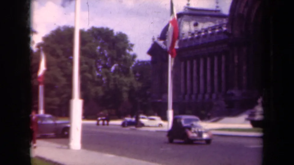 1939 Paris street view with a dark classic car approaching near flagpoles, trees, and the edge of an ornate building.