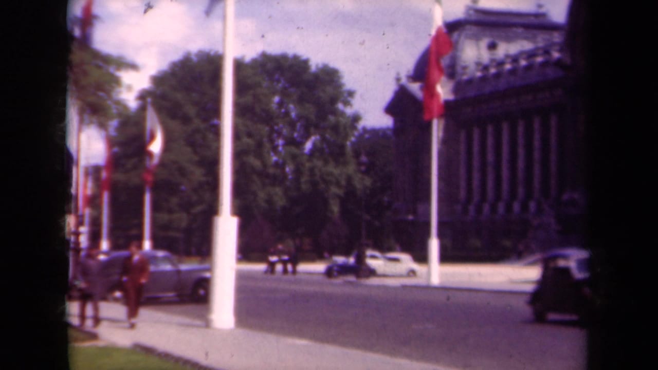 1939 Paris home-movie frame showing French flags along a boulevard, pedestrians on the sidewalk, period cars, trees, and a large columned building.