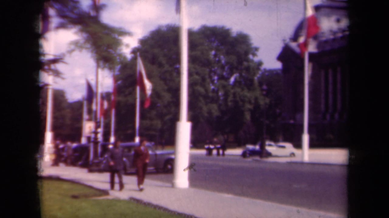 1939 Paris street view with pedestrians in period dress, French flags, cars along the curb, trees, and part of a monumental building.