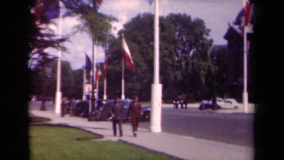 1939 Paris home-movie frame showing two pedestrians approaching the camera near French flags, parked cars, trees, and a broad roadway.