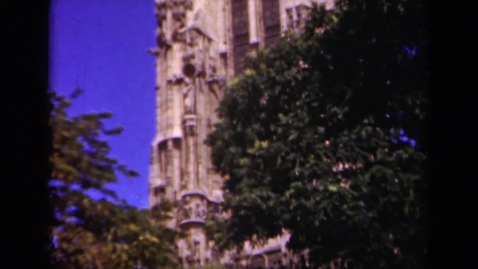 1939 Paris frame showing a Gothic-style stone tower partly hidden by leafy trees, viewed from street level under a bright blue sky.
