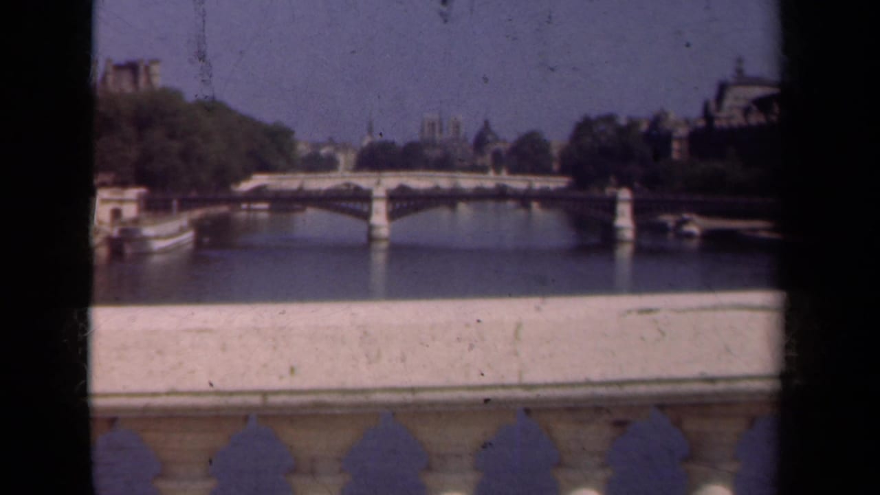 1939 Paris home-movie frame showing a Seine bridge, distant Notre-Dame towers, riverside trees, and a pale stone parapet.
