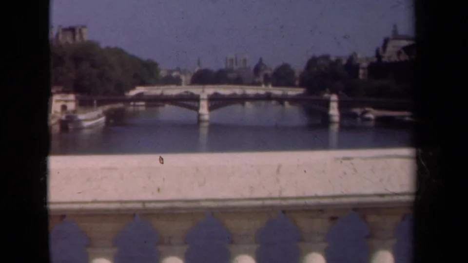 1939 Paris home-movie view of Notre-Dame beyond a Seine bridge, with trees, buildings, and foreground architectural framing.