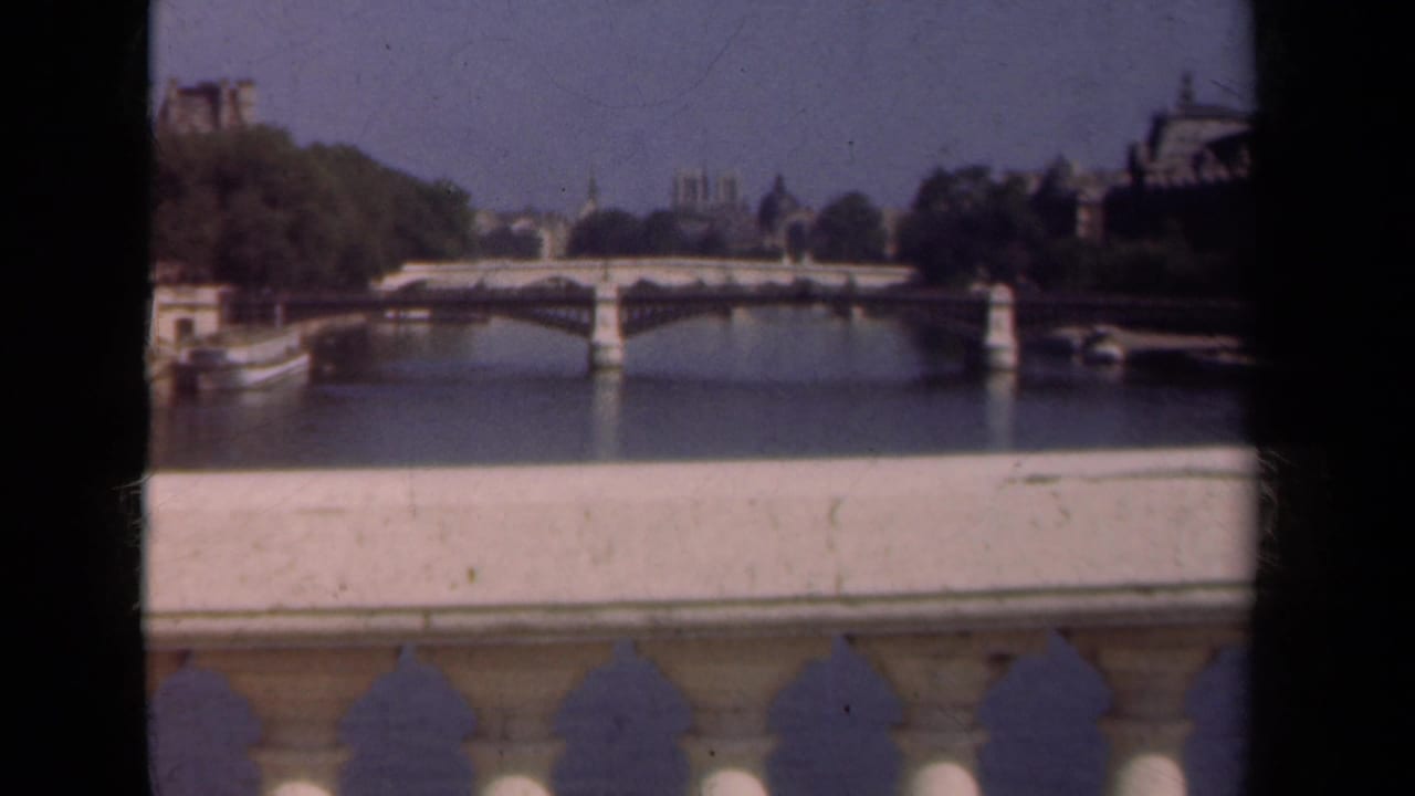 1939 Paris home-movie view across the Seine toward a stone bridge, distant buildings, and a pale bridge balustrade in the foreground.
