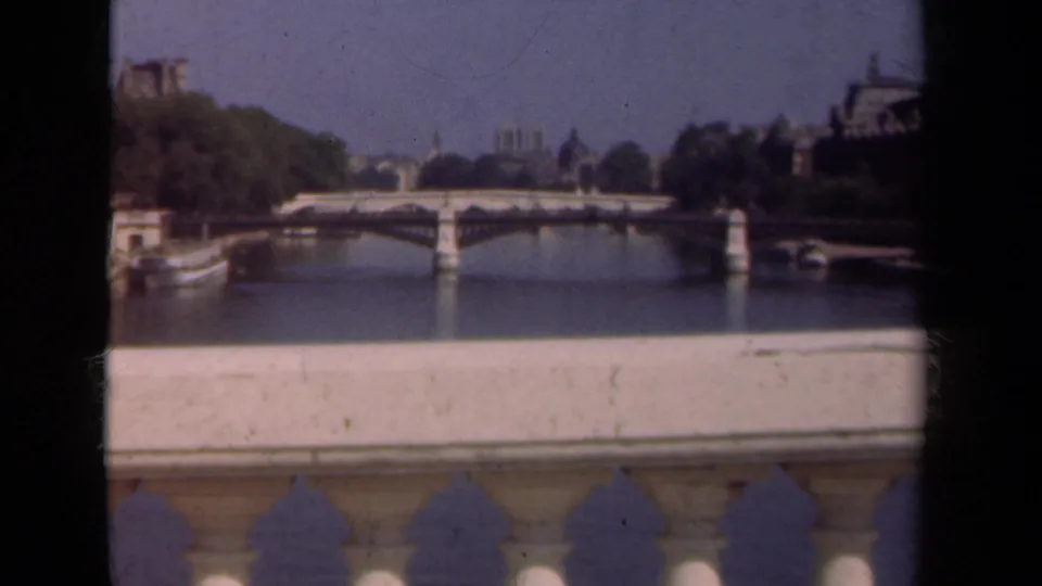 1939 Paris home-movie view across the Seine toward a stone bridge, distant buildings, and a pale bridge balustrade in the foreground.