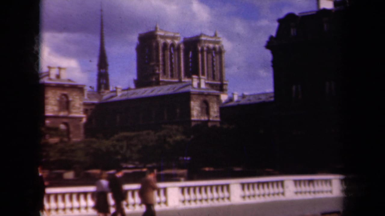 1939 Paris footage showing Notre-Dame Cathedral towers and a nearby spire behind pedestrians walking along a bridge railing.