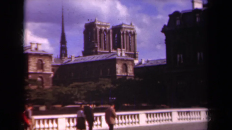 1939 Paris home-movie frame with Notre-Dame, a slender spire, trees, and pedestrians along the bridge in soft color 8mm.