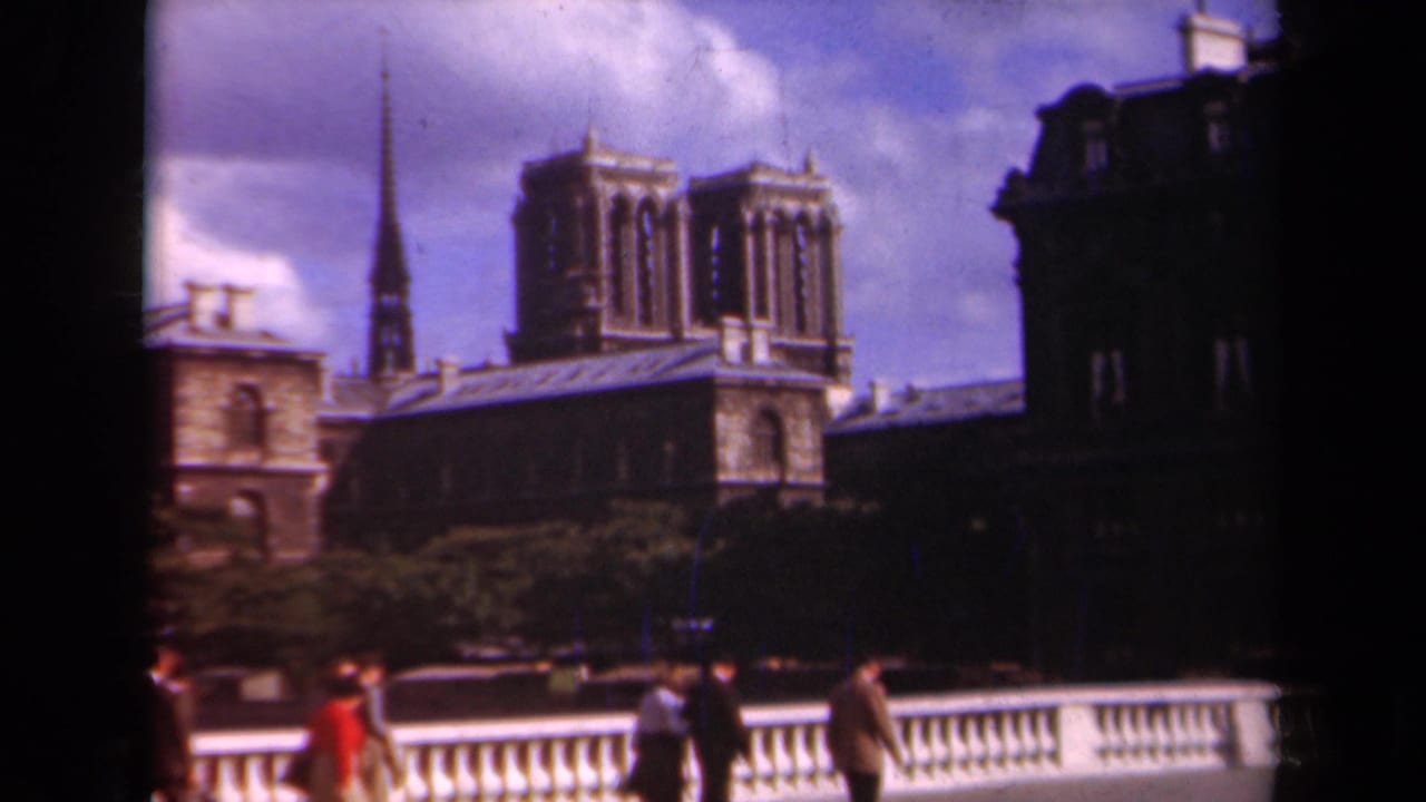 1939 Paris home-movie frame showing Notre-Dame Cathedral, its spire and towers, with pedestrians crossing in front of a bridge railing.