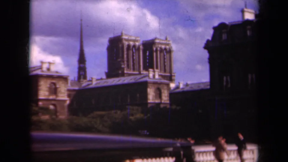 Softly focused 1939 Paris frame showing Notre-Dame Cathedral above trees, with a bridge railing and passing figures near the lower edge.