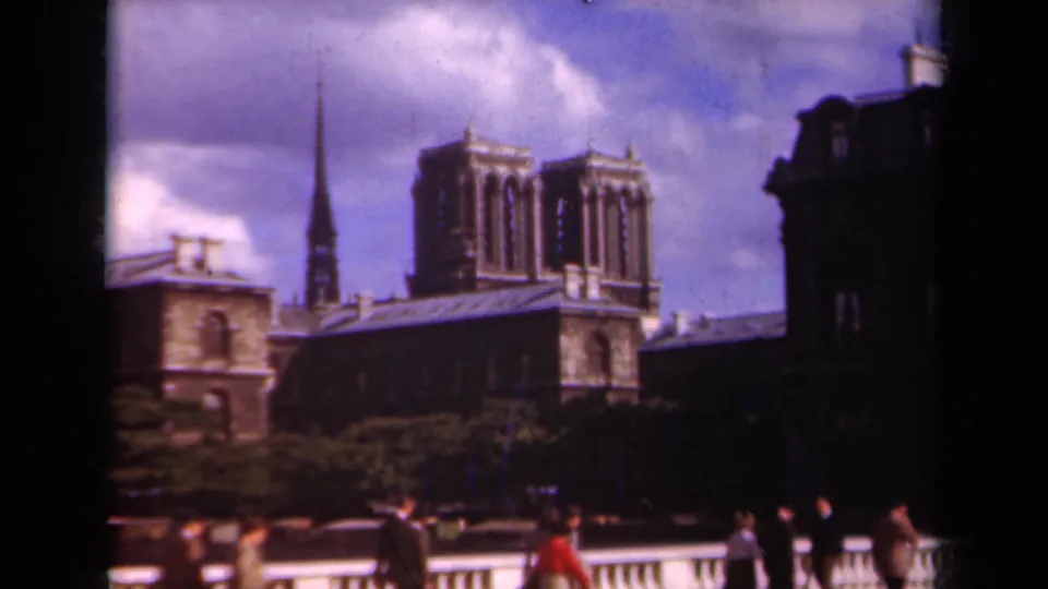 1939 Paris home-movie frame showing Notre-Dame Cathedral behind pedestrians and a bridge balustrade, with clouds and dark edge vignetting.