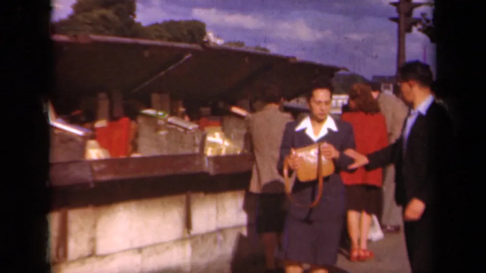 A woman in a dark suit with a tan purse walks past a man beside the 1939 Paris newsstand.