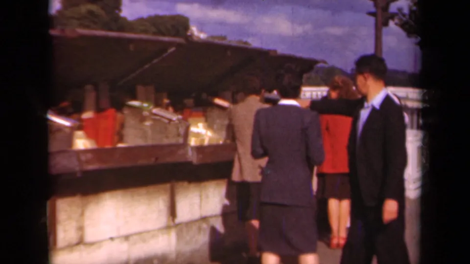 Pedestrians, including a suited man and women in skirts, stand near a Paris newsstand in 1939 footage.