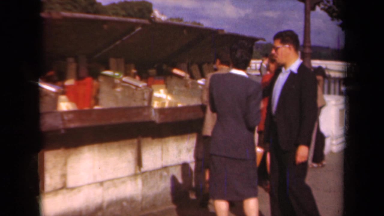 Paris, 1939 footage shows adults in formal dress pausing by a stone market stall with goods displayed under a canopy.
