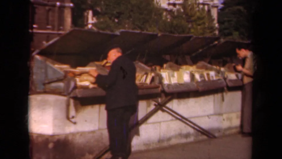 Paris, 1939 footage shows adults leaning over and browsing goods arranged on a long outdoor stall beside a stone wall.