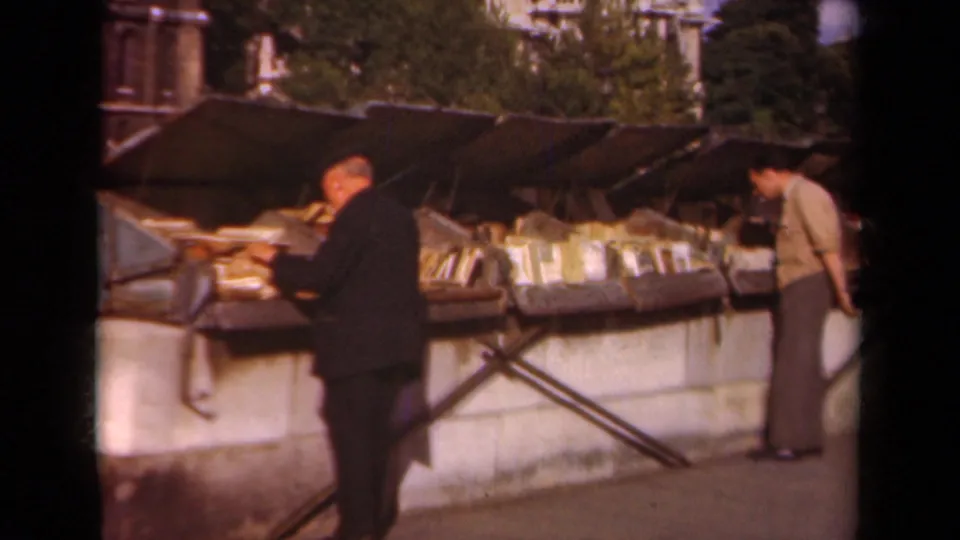 1939 Paris home-movie frame showing men in period clothing browsing books at an outdoor market stall.