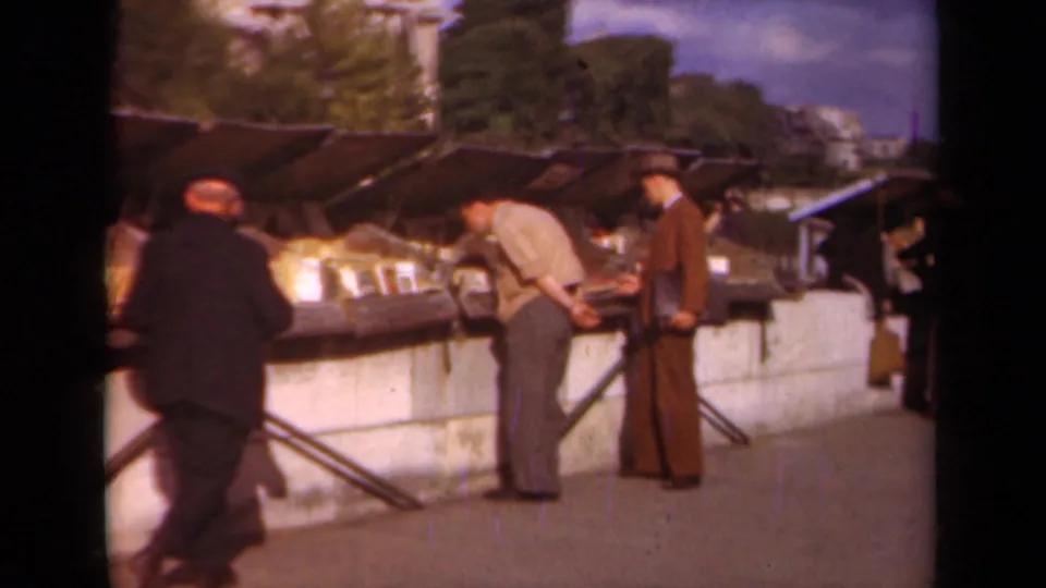 Paris 1939 home-movie frame with suited men examining books at a sunlit outdoor market.