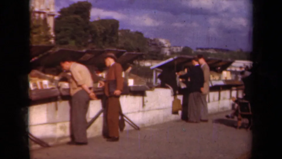 1939 Paris home-movie frame showing suited men browsing awning-covered street-market stalls beside a low wall.