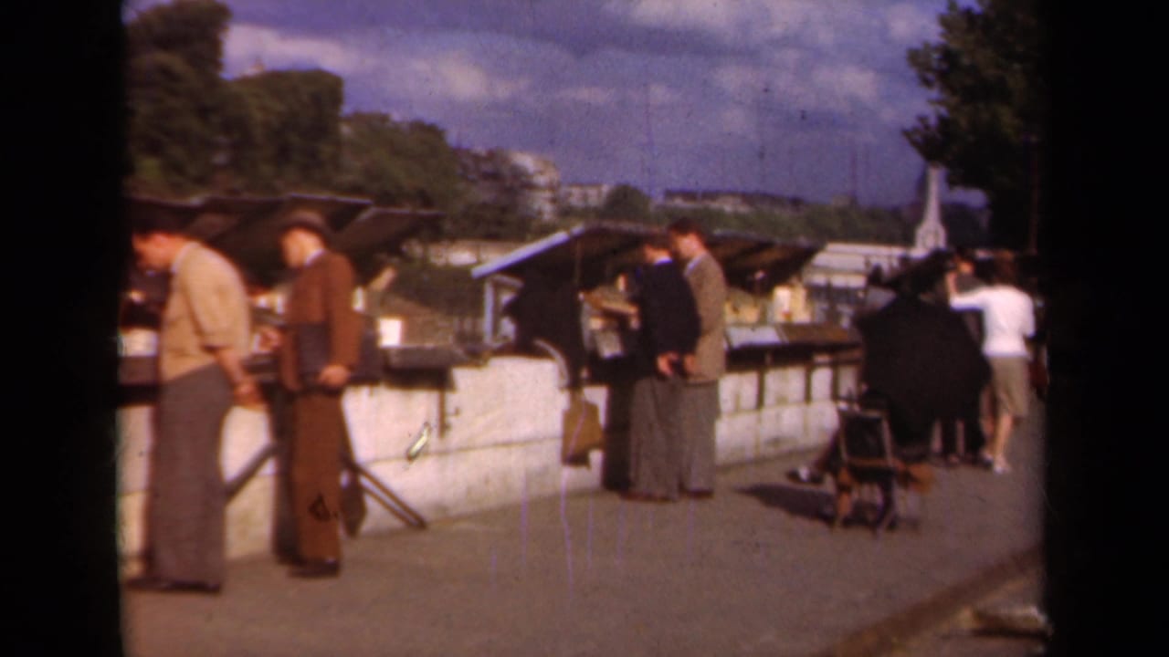 1939 Paris street-market scene with suited men near stalls, a stroller, and pedestrians farther down the promenade.