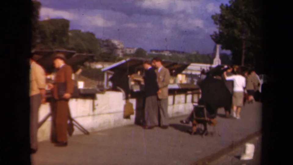 Paris 1939 home-movie frame with men in period suits at market stalls and a woman in a white top walking nearby.