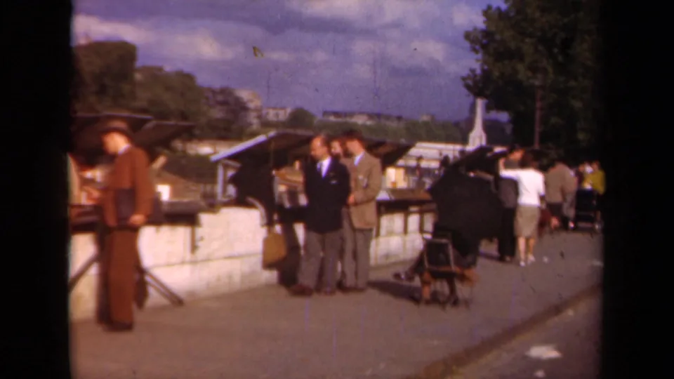 1939 Paris frame showing suited men gathered by outdoor market stalls, with pedestrians and a stroller on the promenade.