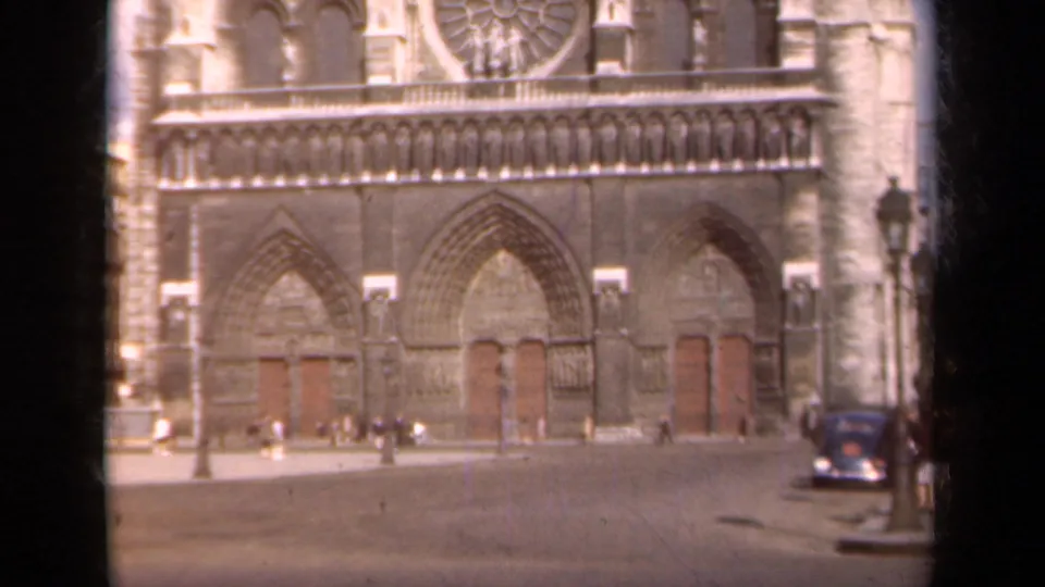 1939 Paris home-movie frame showing Notre Dame's west portals, scattered pedestrians, a lamppost, and a parked dark automobile.