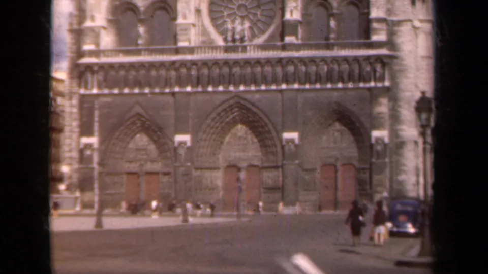 1939 Paris street view of Notre Dame's west front, with people walking across the plaza and a dark automobile at the right edge.