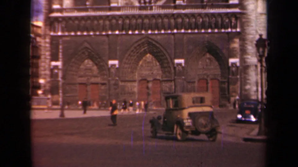 Notre Dame Cathedral exterior in Paris as pedestrians gather near the portals and a light-colored period car passes through the foreground.