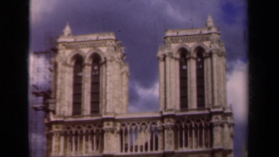 Paris 1939 home-movie frame of Notre-Dame's towered facade, with Gothic stone details, blue sky, clouds, and scaffolding beside the structure.