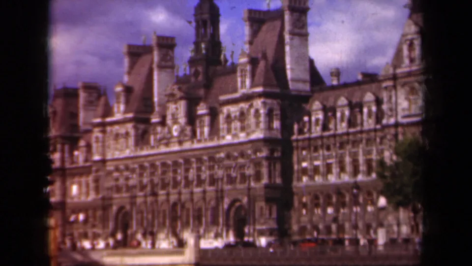 1939 Paris frame showing the Hôtel de Ville exterior, mansard roofs, arched windows, trees, cloudy sky, and a small red vehicle at street level.