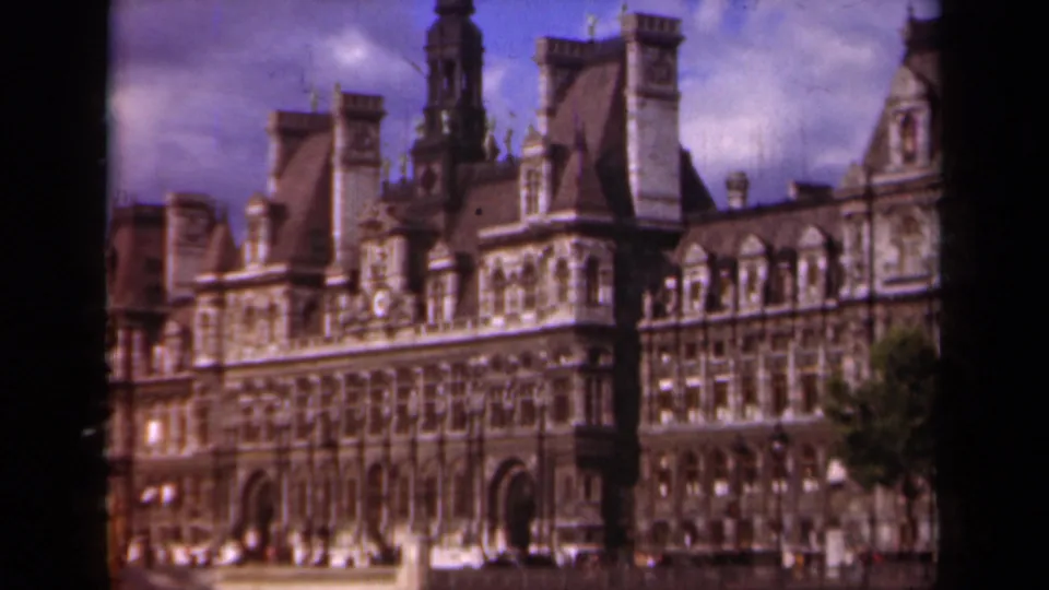 Soft 8mm frame of the Hôtel de Ville in Paris, showing arched entrances, decorative stonework, roof towers, and cloudy sky.
