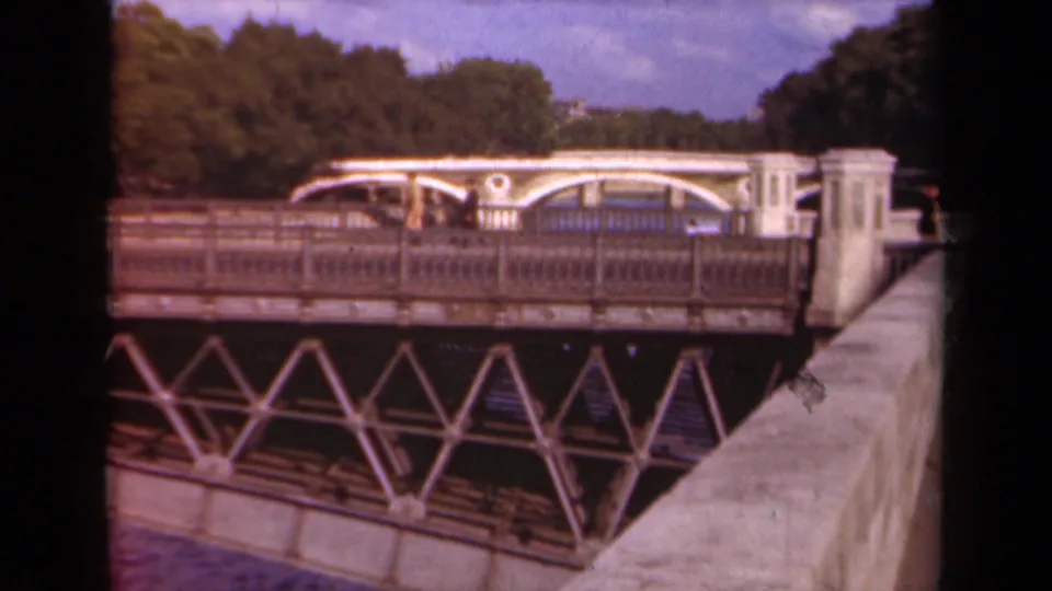 1939 Paris 8mm frame of a metal bridge and riverside wall, with small pedestrians crossing and a tree-lined background.