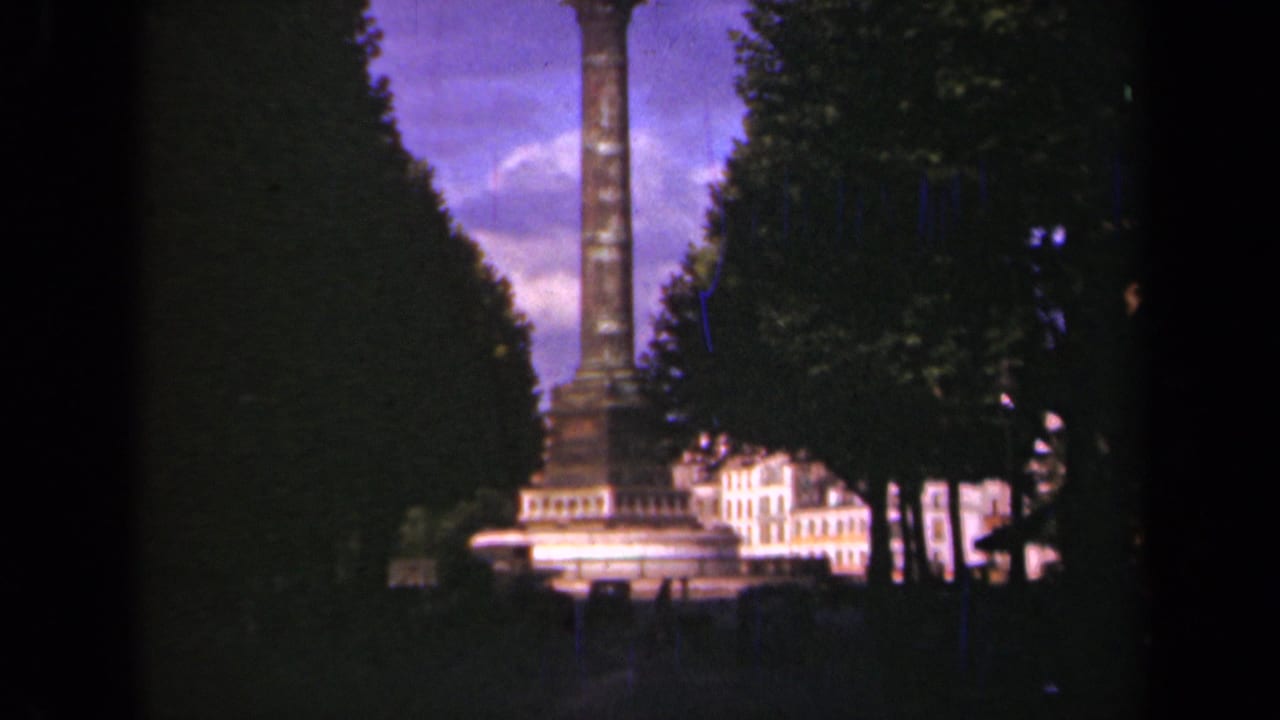 1939 Paris home-movie frame showing a tall column monument between tree canopies with pale buildings behind it.