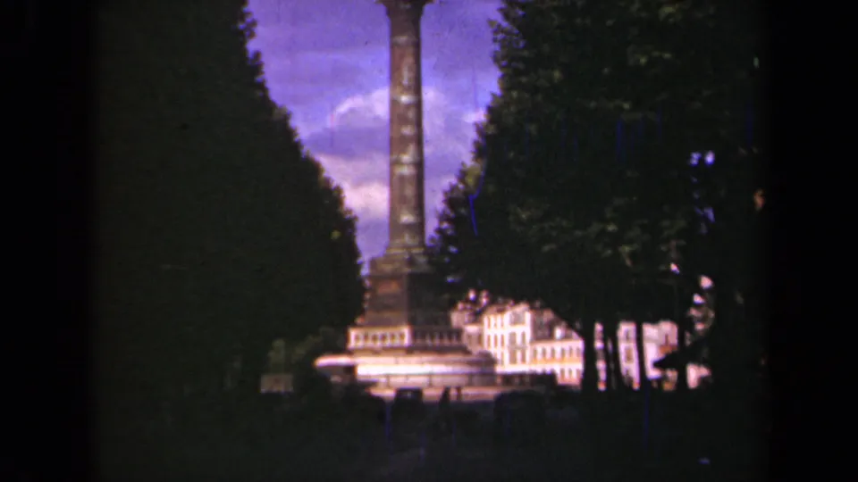 1939 Paris home-movie frame showing a tall column monument between tree canopies with pale buildings behind it.