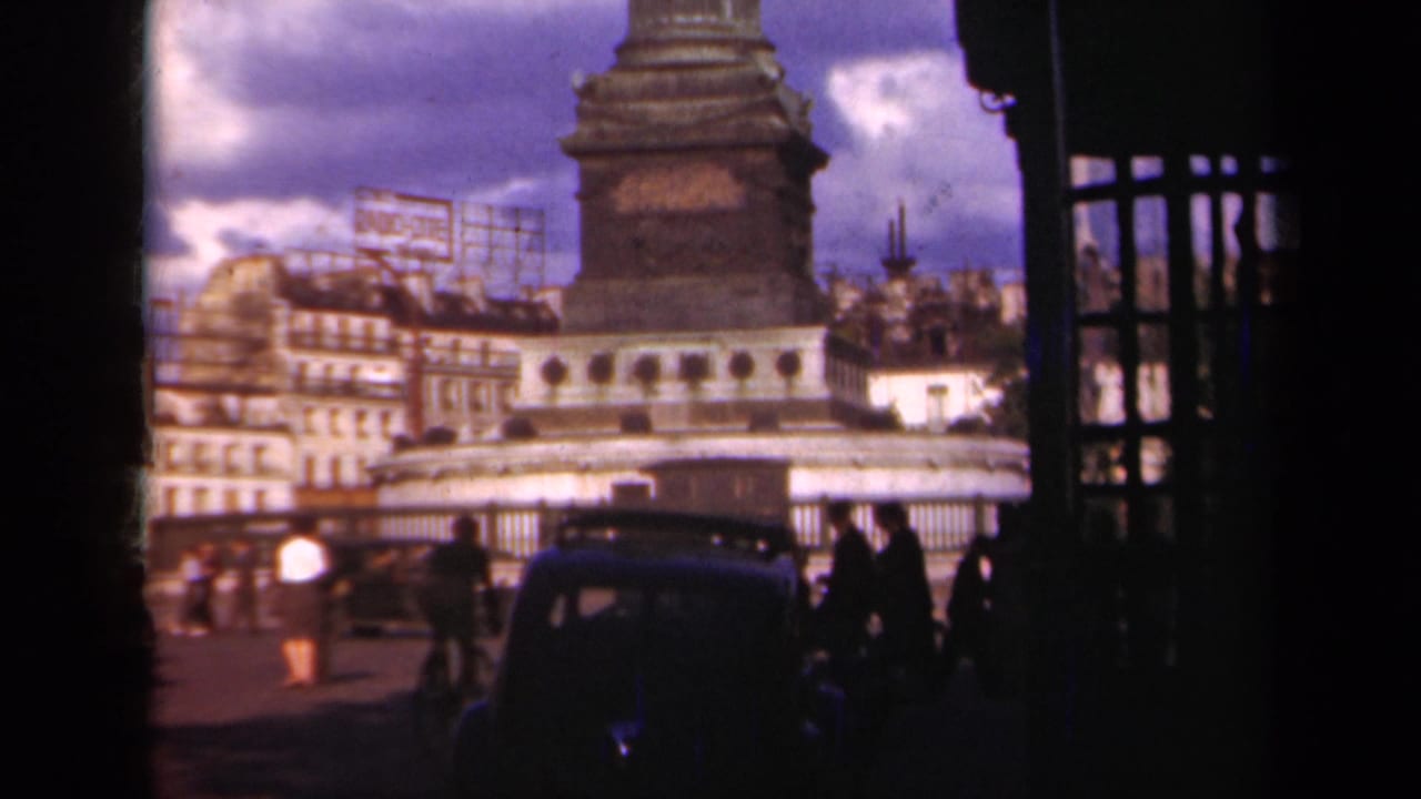 1939 Paris street-level home-movie frame showing the base of a column monument, pedestrians, older cars, railings, buildings, and rooftop sign frames.