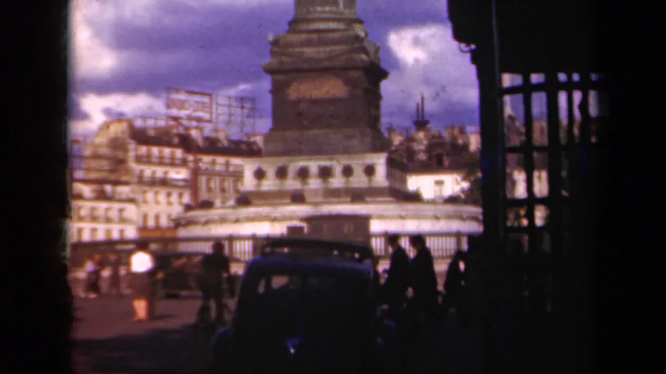 1939 Paris street-level home-movie frame showing the base of a column monument, pedestrians, older cars, railings, buildings, and rooftop sign frames.