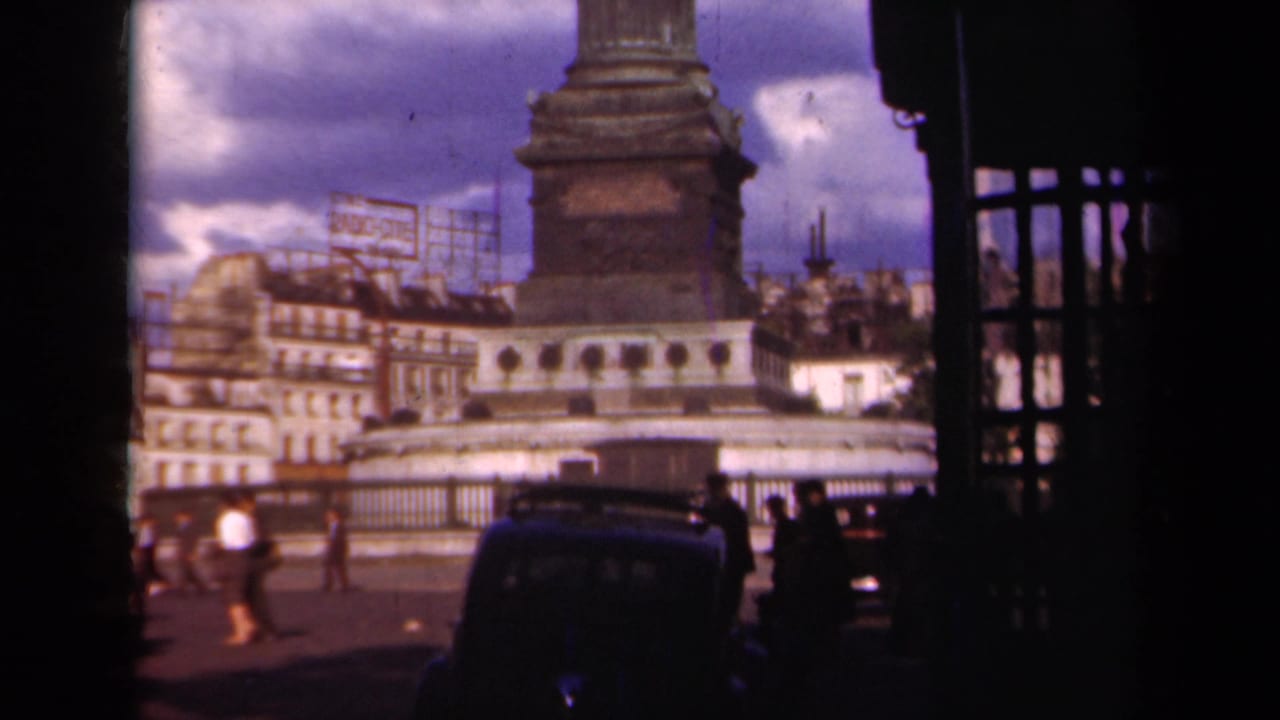 Paris, 1939 home-movie frame with the monument base above a street scene of pedestrians, classic cars, railings, buildings, and rooftop signage.