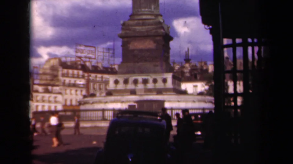 Paris, 1939 home-movie frame with the monument base above a street scene of pedestrians, classic cars, railings, buildings, and rooftop signage.