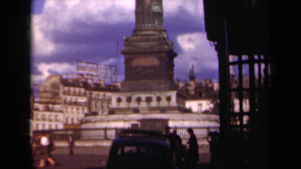 1939 Paris home-movie frame showing the base of a monumental column with pedestrians, buildings, a car, and dark framing in the foreground.