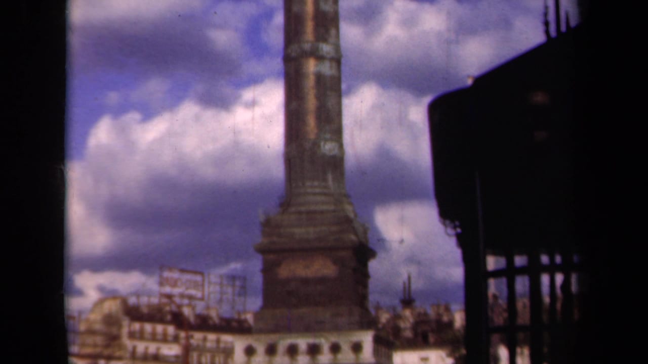 Column base and lower shaft seen between dark vertical framing with buildings and signage nearby.
