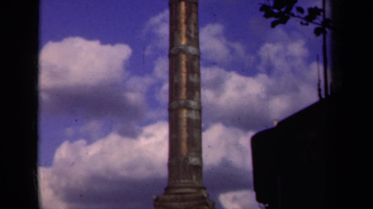 1939 Paris home-movie frame looking upward along the column shaft against blue sky, clouds, and dark foreground silhouettes.