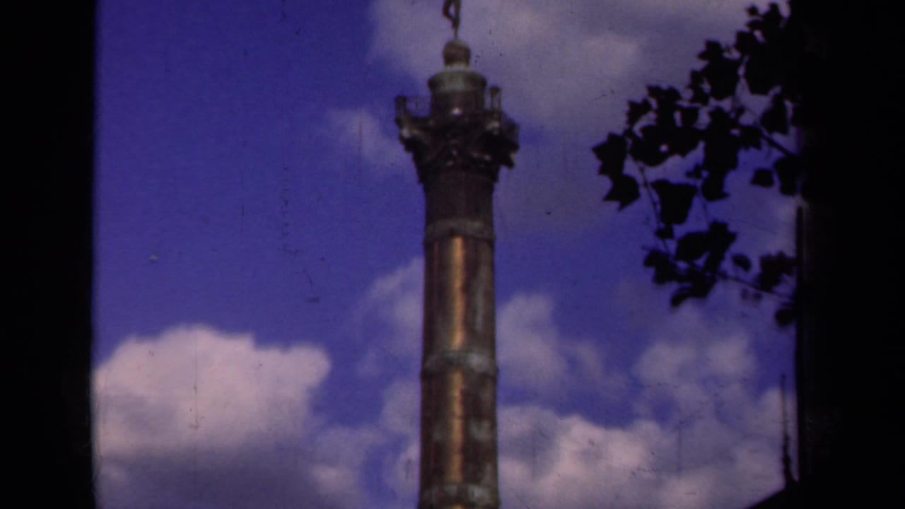 Upward view of a bronze column topped by a winged statue against blue sky and clouds.