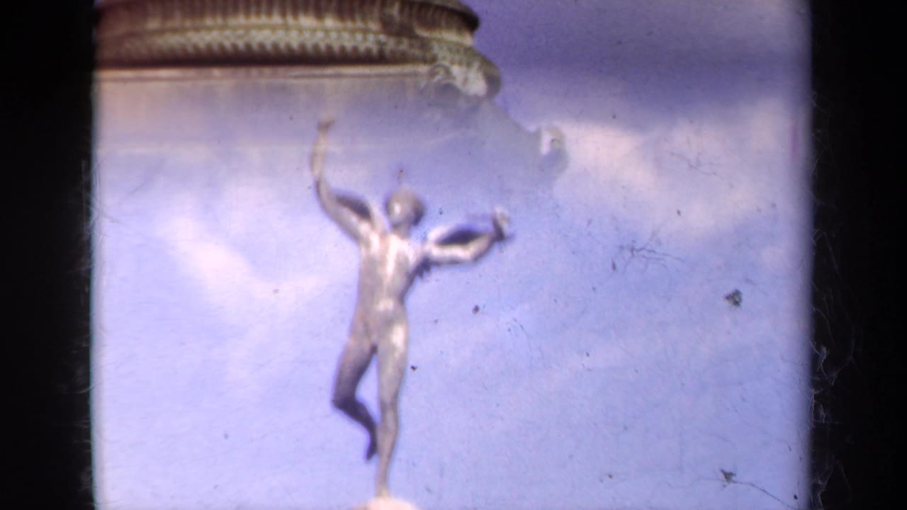 1939 Paris 8mm frame showing a winged figure statue above the monument, filmed upward against a pale blue and cloudy sky.