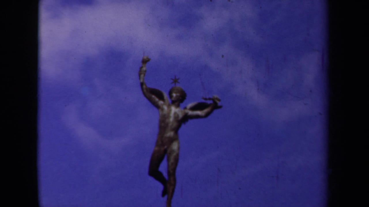 1939 Paris home-movie still of a bronze winged figure beneath drifting clouds, with visible grain and color shift.